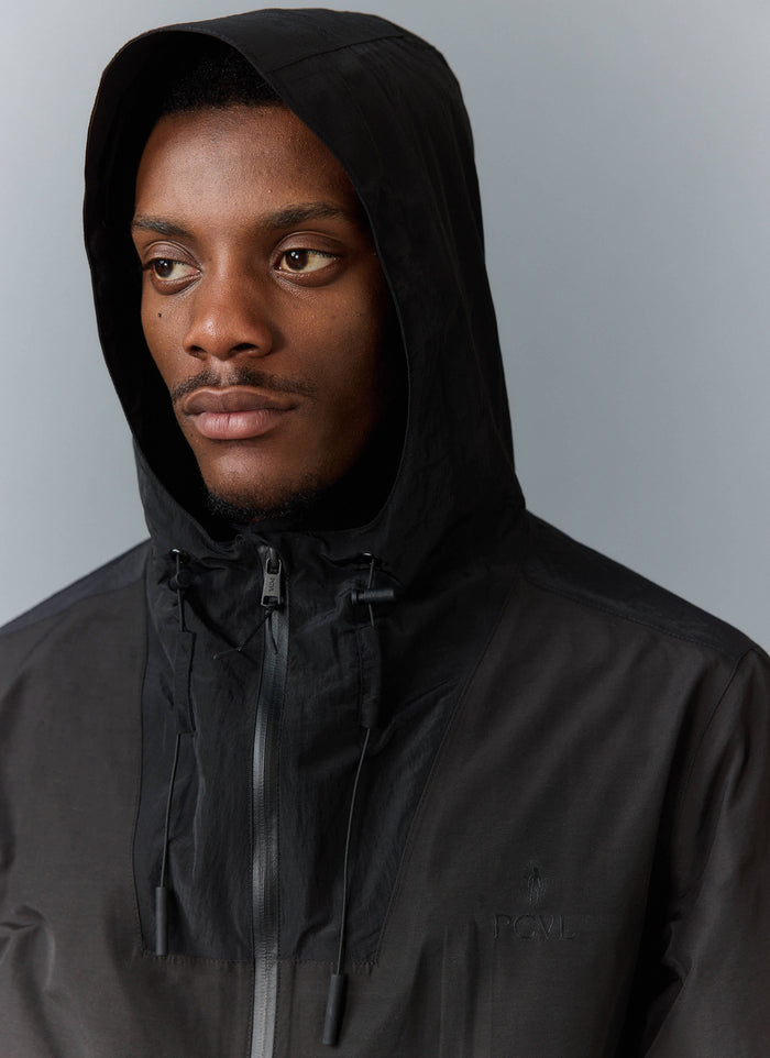 Close-up portrait of a model wearing the black PCVL Zip Through Water Resistant Jacket from Percival with the hood up.
