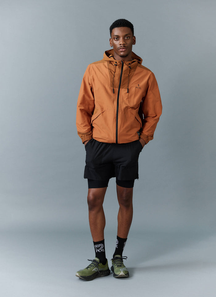 A model wears the rust orange PCVL Zip Through Water Resistant Jacket from Percival with black shorts and PCVL socks.