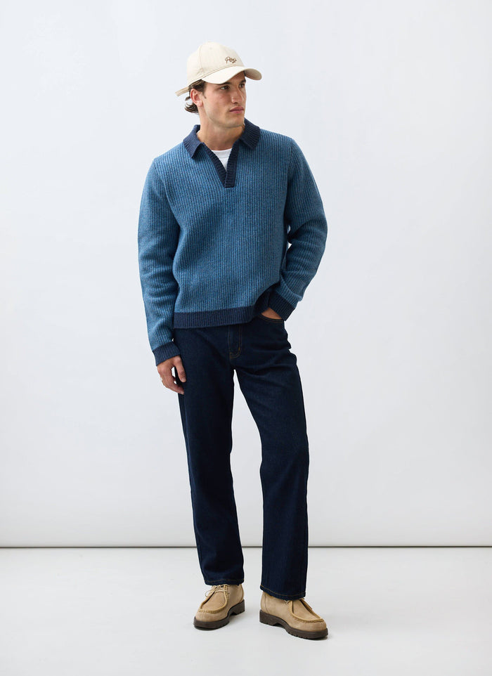 A man models the Percival Picnic Polo in two-toned blue, a ribbed cotton knit, styled with dark jeans and a baseball cap.