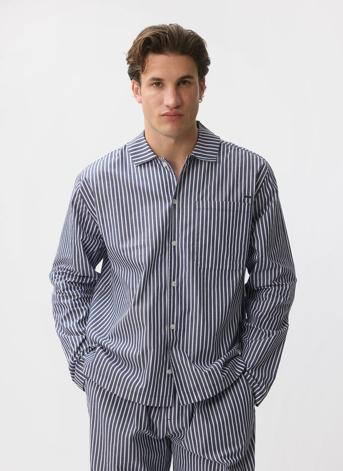 A model wears the Percival pyjama set in a blue and white cotton stripe, standing against a clean white studio background.