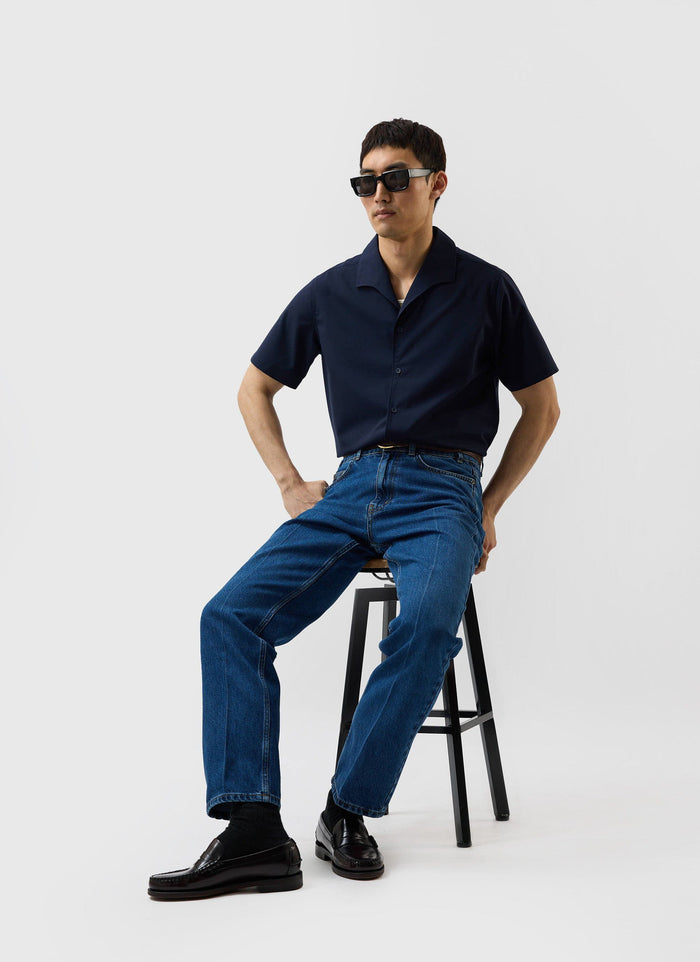 A model sits on a stool wearing the navy blue Percival x Warren Relaxed Cuban Drape Shirt with jeans and sunglasses.