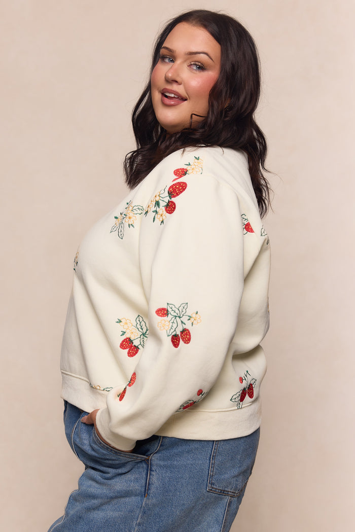 A woman with long dark hair smiles softly, posing sideways in the Ivy City Co Sabina Sweatshirt in Cream, a cotton fleece maternity top with colorful embroidered strawberries and flowers, paired with blue jeans against a neutral background.