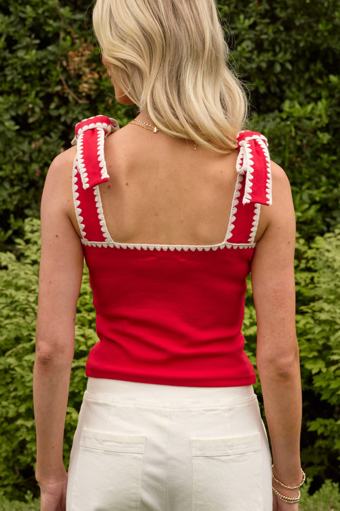 A woman with blonde hair, seen from behind, wears the Ivy City Co Sammi Top in Firework Red with white trim and bows—perfect for the Fourth of July—paired with white pants and standing outdoors in front of green bushes.