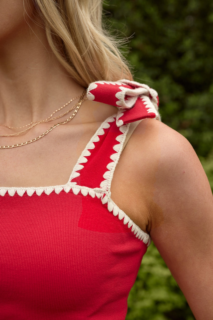 Close-up of a person wearing the Ivy City Co Sammi Top in Firework Red, featuring cream trim and a shoulder bow—perfect for the Fourth of July—styled with layered gold necklaces against a softly blurred green backdrop.