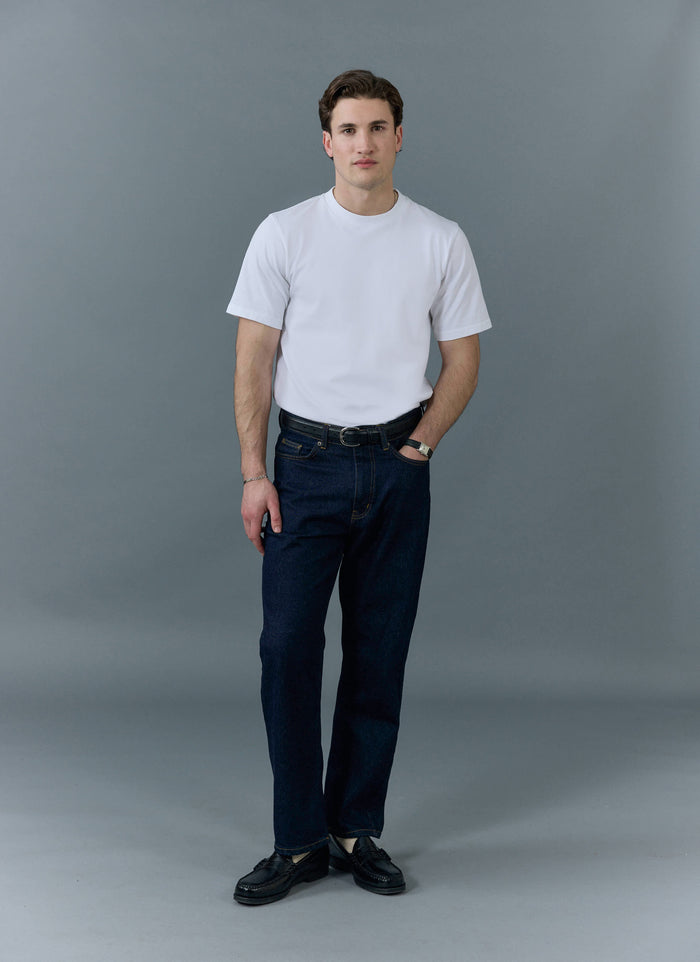 A man models the Percival Straight Leg Denim Jeans in dark indigo, styled with a white t-shirt and black loafers.