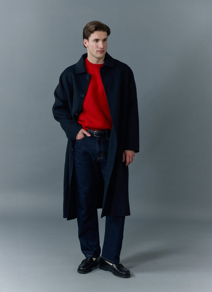 A model poses in a studio wearing the Percival dark indigo straight leg jeans, styled with a red jumper and black overcoat.