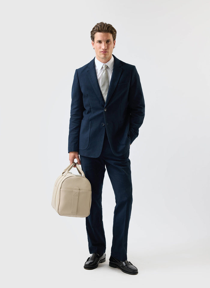 A man models the Percival Tailored Moleskin Blazer in navy, styled as a full suit and carrying a cream weekend bag.