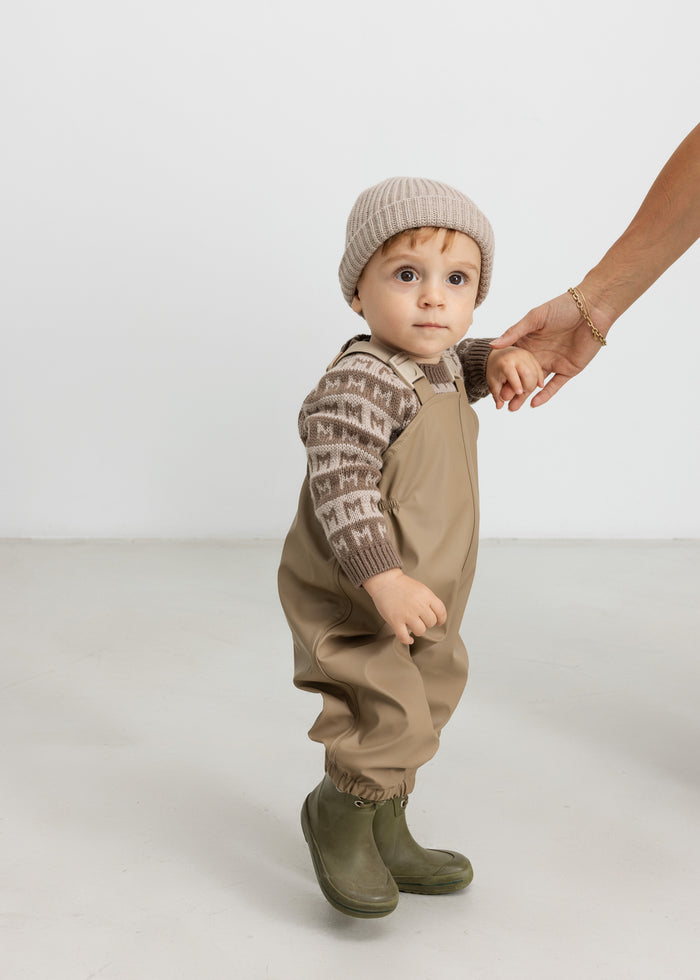 Baby wearing brown M-patterned sweater, thermal pants and hat, holding an adult's hand