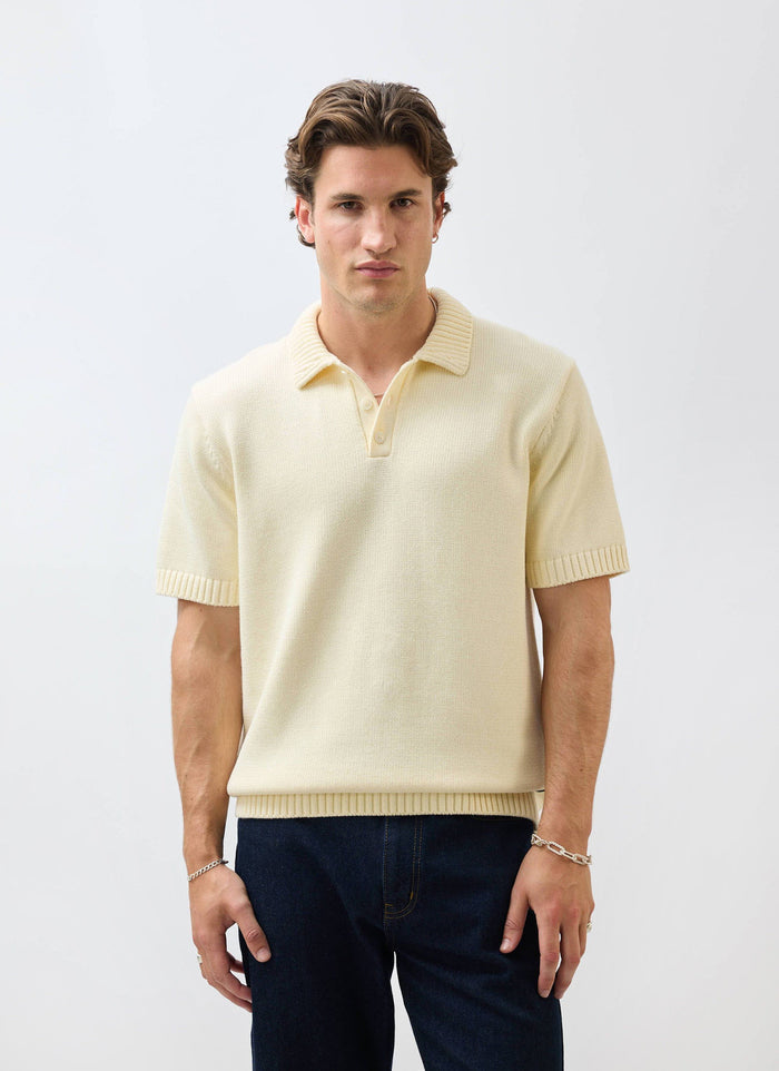 A man models the Percival Verso Short Sleeve Knitted Shirt in ecru, styled with dark jeans against a white background.