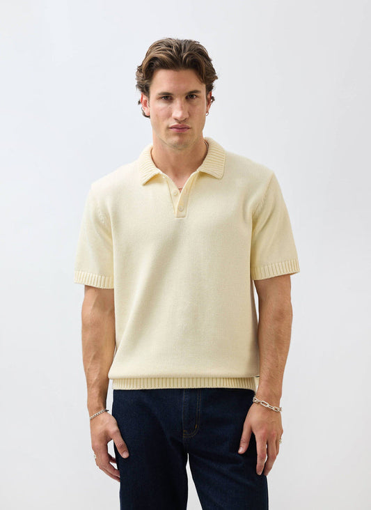 A man models the Percival Verso Short Sleeve Knitted Shirt in ecru, styled with dark jeans against a white background.