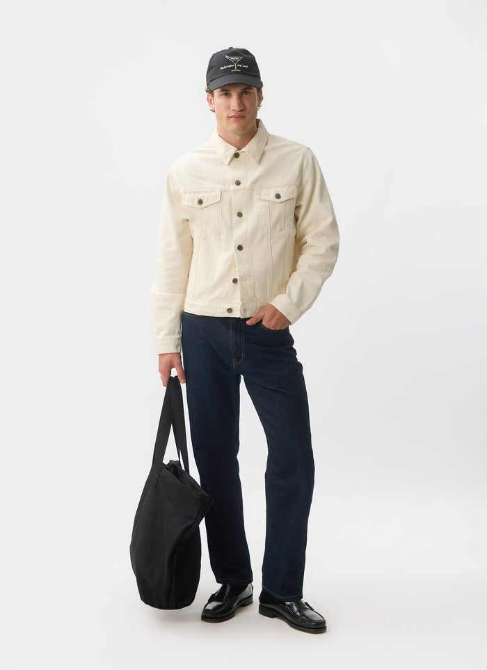 A full-length shot of a model wearing the ecru denim Western Trucker Jacket from Percival, styled with dark jeans and a cap.