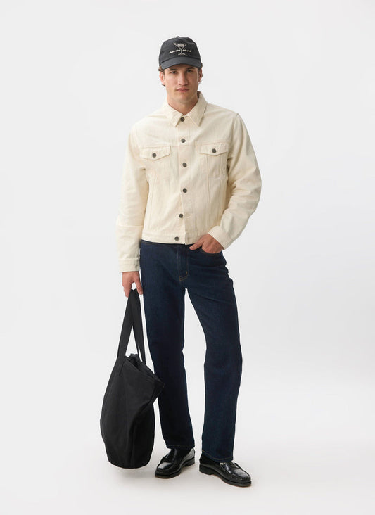 A full-length shot of a model wearing the ecru denim Western Trucker Jacket from Percival, styled with dark jeans and a cap.