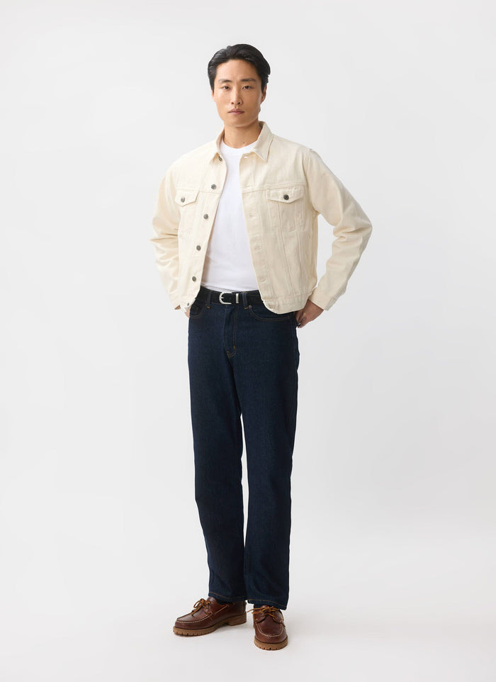 A model wears the Percival Western Trucker Jacket in ecru denim with a white t-shirt, dark jeans, and brown shoes.