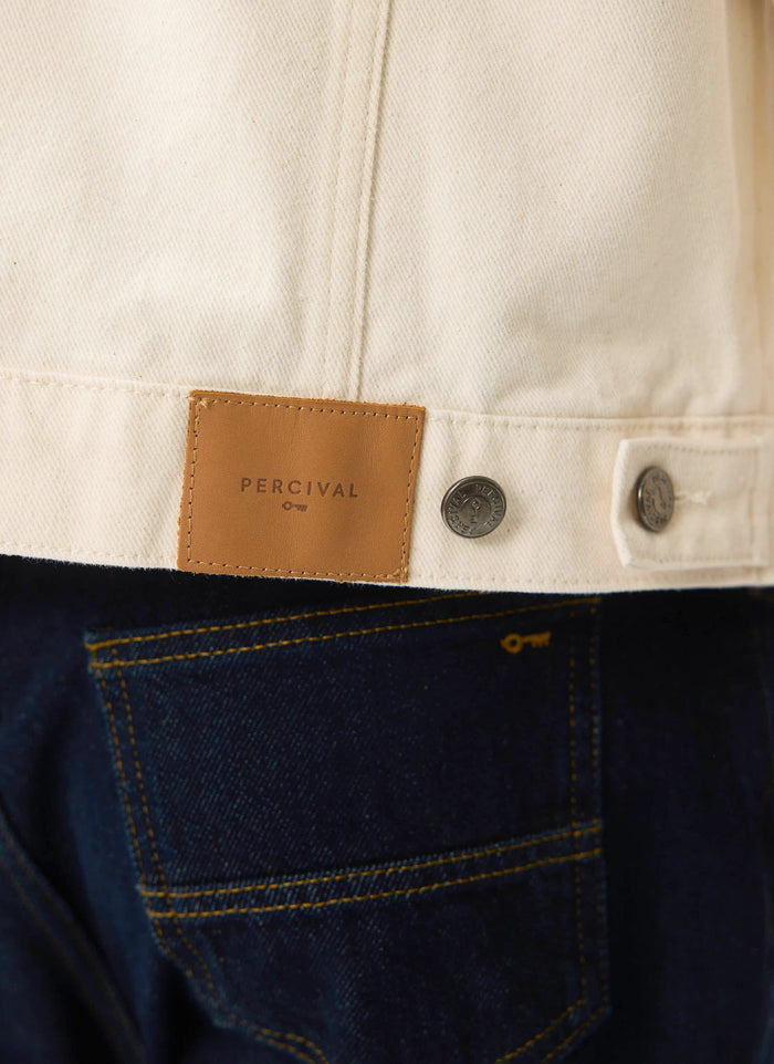 Close-up of the brown leather Percival patch on the back of the ecru Western Trucker Jacket, worn over dark denim jeans.