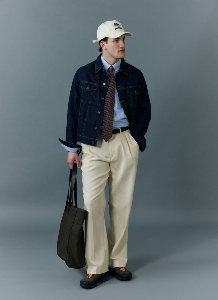 A man models the Percival Western Trucker Jacket in Dark Indigo, styled over a shirt and tie with cream trousers.