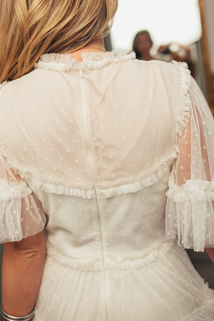 The Whimsical Dress in White by Ivy City Co features a sheer, polka-dotted maxi design with ruffles, lace, and short puffed sleeves. A tulle skirt and loose blonde hair make the look effortlessly dreamy from the back view.