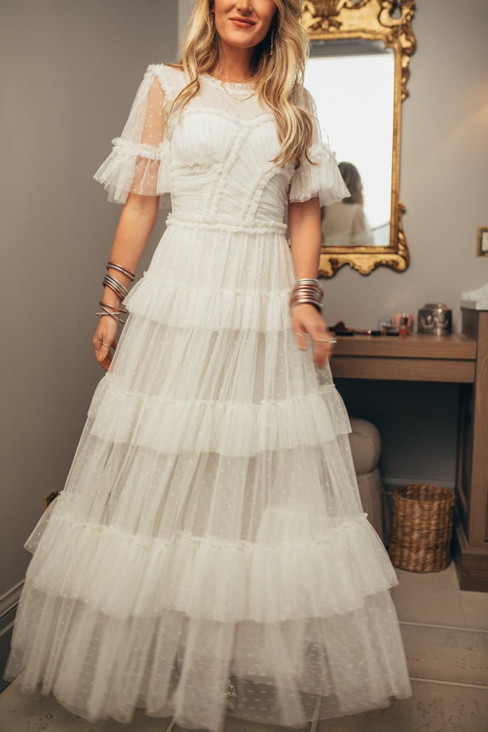 A woman wearing the Ivy City Co Whimsical Dress in White—a layered tulle maxi dress with sheer sleeves—stands in a room adorned with a gold mirror, vanity table, and woven basket. She accessorizes with multiple bangles on her wrist and wears her loose, wavy blonde hair for an effortlessly whimsical appearance.